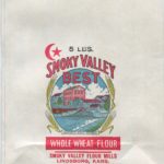 A large, flat unused flour bag in full color. At the center in an oval frame is a view of a red brick, three-story mill building with a gabled roof, surrounded by lower level industrial buildings. In the foreground is a blue river that has been dammed. On either side of the frame are stalks of yellow wheat. The label at the bottom reads "Smoky Hill River." To the upper left is a red crescent moon and star. Text on the bag reads: "5 pounds. Smoky Valley Best. Whole Wheat Flour. Smoky Valley Flour Mills. Lindsborg, Kansas."