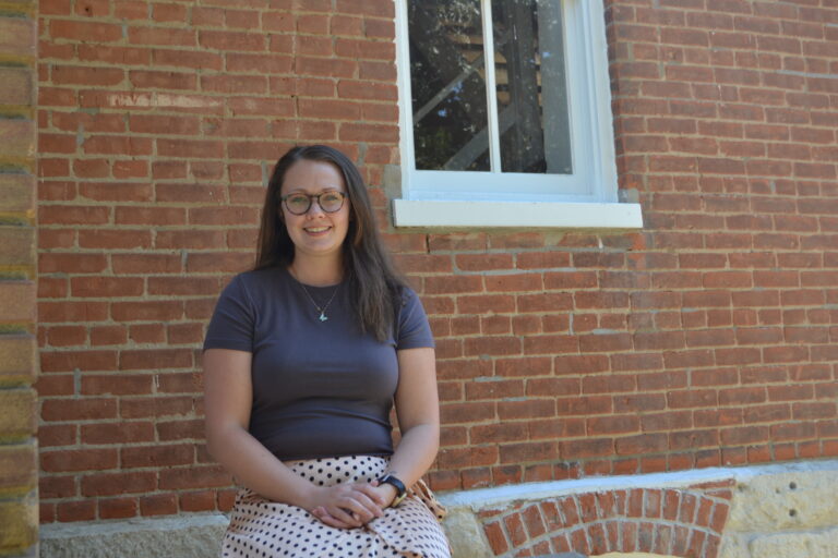 Aubrey Wheeler - a woman with glasses seated in front of a brick wall outside.